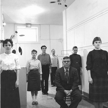 A black and white photo of a man sitting in a chair while 6 people stand scattered behind him
